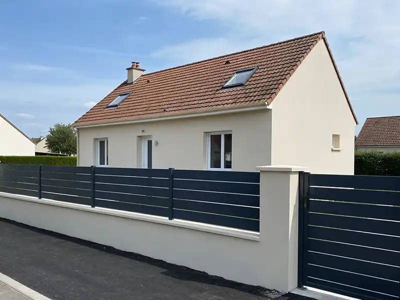 Une petite maison moderne de couleur cr&egrave;me avec un toit de tuiles rouges, des fen&ecirc;tres &agrave; cadre blanc et des lucarnes. La propri&eacute;t&eacute; est dot&eacute;e d'une cl&ocirc;ture et d'un portail en lattes horizontales gris fonc&eacute;, issus d'un projet de cr&eacute;ation muret et cloture, et d'un trottoir qui longe la fa&ccedil;ade.