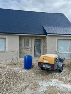 Une maison nouvellement construite avec un enduit ext&eacute;rieur, un toit sombre et des fen&ecirc;tres couvertes, entour&eacute;e de tas de gravier et de mat&eacute;riel de construction, dont un tonneau bleu et un compresseur d'air jaune sur une remorque &agrave; l'avant.