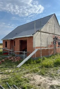 Une maison en construction avec des &eacute;chafaudages sur le c&ocirc;t&eacute;, des murs ext&eacute;rieurs partiellement finis en attente d'enduit ext&eacute;rieur, des briques apparentes, des mat&eacute;riaux de construction et de l'herbe au premier plan sous un ciel partiellement nuageux.