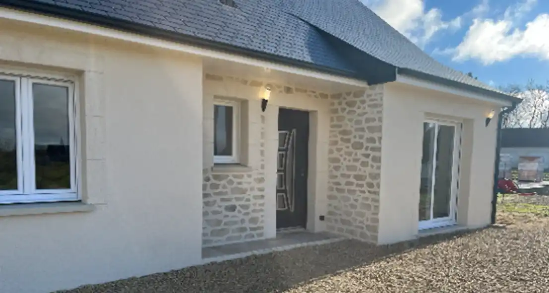 Maison de plain-pied avec une fa&ccedil;ade ext&eacute;rieure en pierre claire et enduit cr&egrave;me, un toit sombre, une porte d'entr&eacute;e moderne, deux fen&ecirc;tres et une cour en gravier. Le ciel est partiellement nuageux &agrave; l'arri&egrave;re-plan.