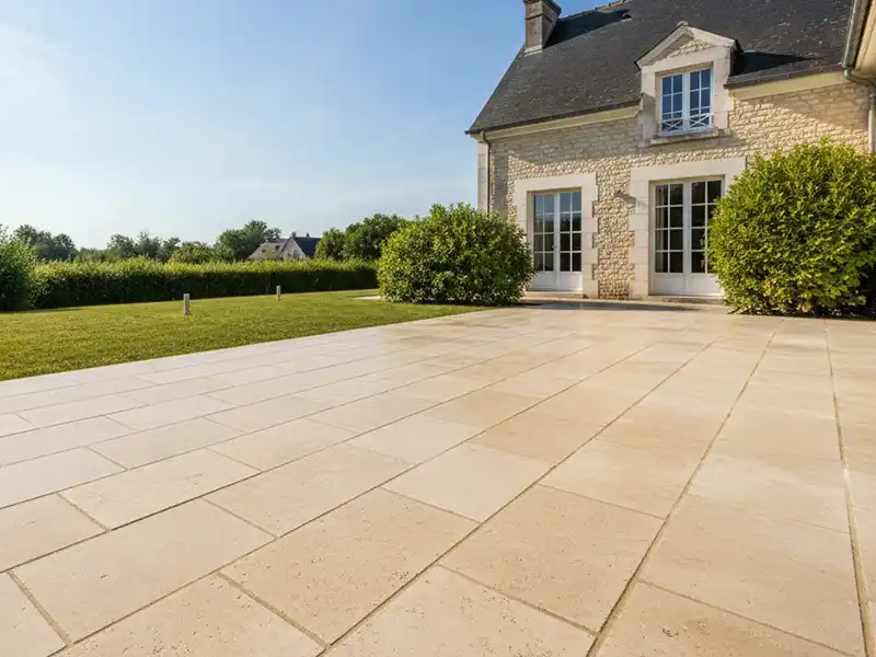 Un grand patio carrel&eacute; de couleur claire &agrave; l'ext&eacute;rieur d'une maison en pierre avec des fen&ecirc;tres et des portes &agrave; cadre blanc, entour&eacute; de buissons verts et d'une pelouse bien entretenue sous un ciel bleu clair, id&eacute;al pour mettre en valeur un nettoyage dallage expert.