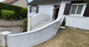 Une personne se tient debout sur un escabeau pour peindre l'habillage ext&eacute;rieur d'une maison blanche aux volets bruns, qui poss&egrave;de un escalier courbe menant &agrave; l'entr&eacute;e et &agrave; un petit jardin.
