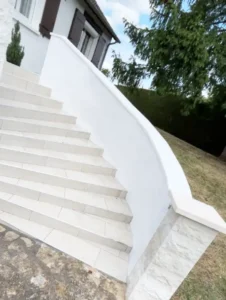 Un escalier ext&eacute;rieur carrel&eacute; blanc avec une rampe blanche incurv&eacute;e et un escalier propre m&egrave;ne &agrave; une maison aux volets bruns, entour&eacute;e de gazon, d'arbres et d'un sentier en pierre.