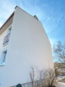 Une maison de trois &eacute;tages avec un mur lat&eacute;ral blanc, plat et sans fen&ecirc;tre, r&eacute;cemment mis en valeur par la r&eacute;novation de la fa&ccedil;ade, quelques arbustes nus &agrave; la base, et un ciel bleu vif au-dessus.