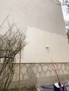 Une personne utilise un rouleau de peinture sur une perche pour appliquer de la peinture de fa&ccedil;ade sur le mur ext&eacute;rieur brut d'un b&acirc;timent, cr&eacute;ant ainsi un contraste &eacute;vident entre les zones fra&icirc;chement peintes et les zones non peintes. Un buisson sans feuilles se trouve &agrave; proximit&eacute;.